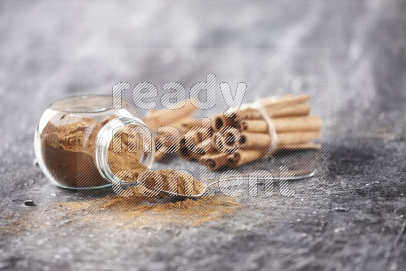Herbal glass jar full cinnamon powder flipped and a metal spoon full of powder, cinnamon sticks stacked and bounded in the back on textured black background in different angles