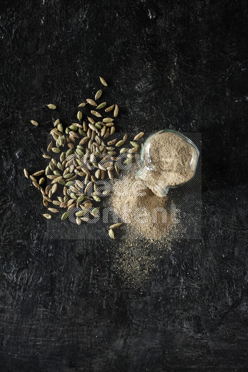 A flipped glass spice jar full of cardamom powder and powder spilled out of it with cardamom seeds spreaded on textured black flooring