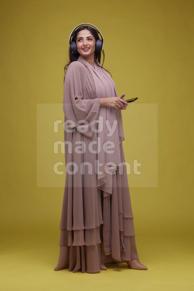 A Saudi woman Listing to Music on a Yellow Background wearing Brown Abaya