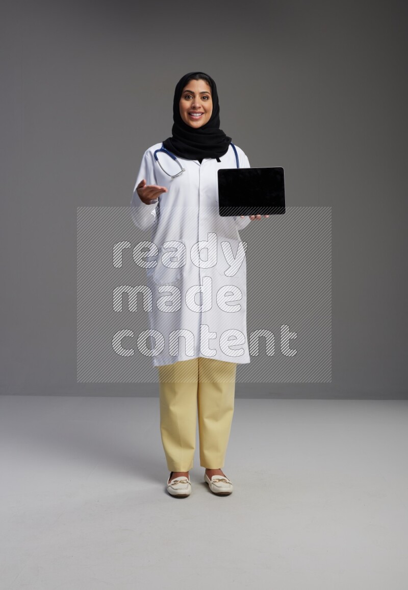 Saudi woman wearing lab coat with stethoscope standing showing tablet to camera with sign in the back on Gray background