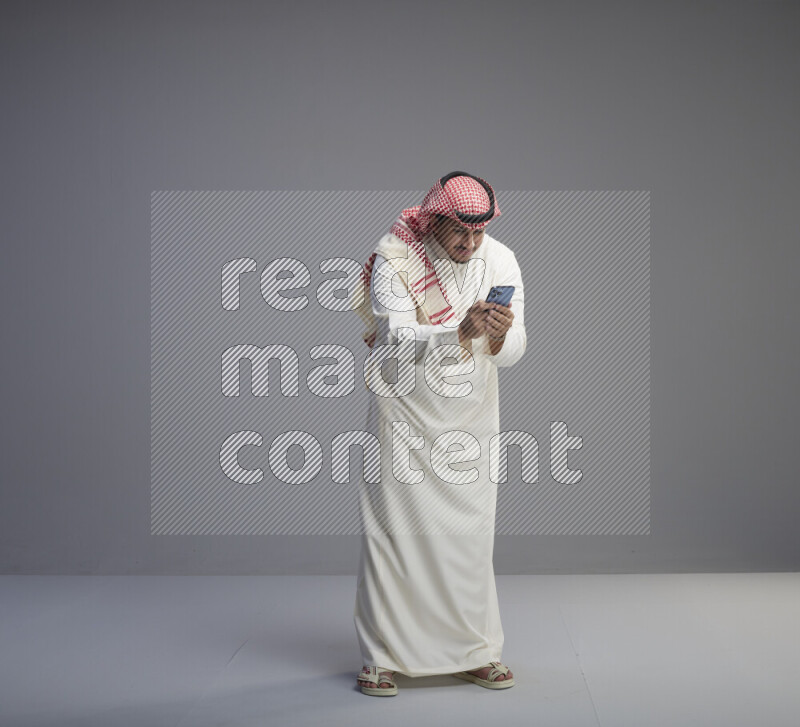 A  Saudi man standing wearing thob and red shomag texting on phone on gray background
