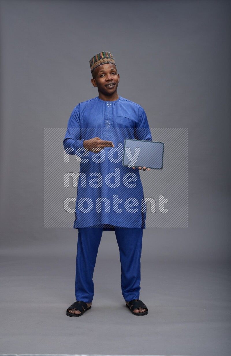Man wearing Nigerian outfit standing showing tablet to camera on gray background