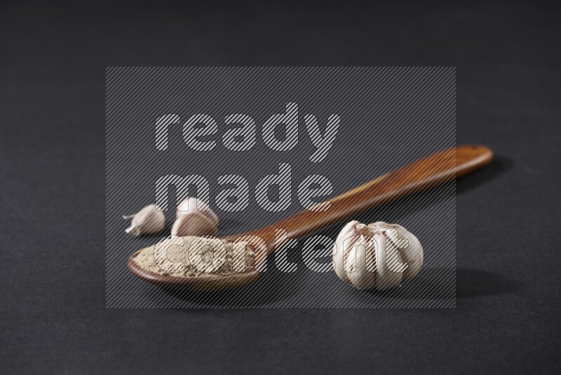 A wooden ladle full of garlic powder with a whole garlic bulb and some cloves beside it on a black flooring