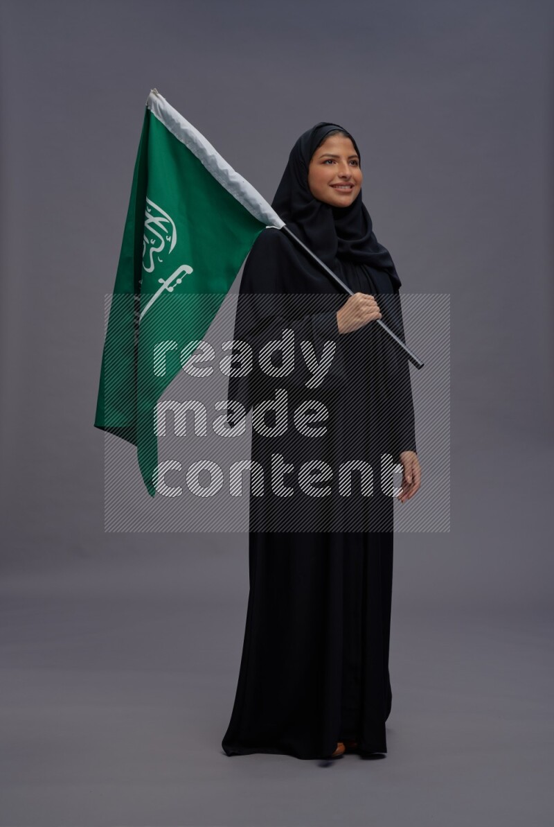 Saudi woman wearing Abaya standing holding Saudi flag on gray background