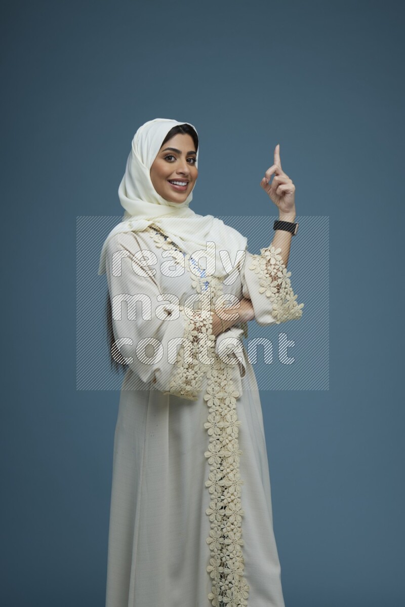 A Saudi woman pointing in a blue background wearing an off-white Abaya Hijab