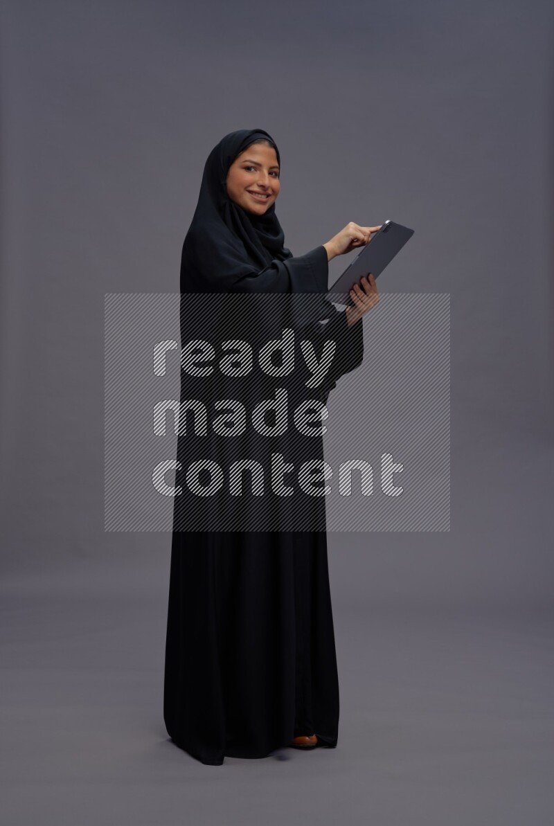 Saudi woman wearing Abaya standing working on tablet on gray background