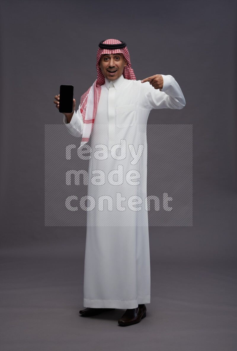 Saudi man wearing thob and shomag standing showing phone to camera on gray background