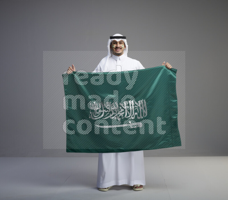 A Saudi man standing wearing thob and white shomag with face painting holding big Saudi flag on gray background