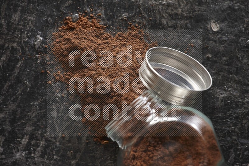 A flipped glass spice jar full of cloves powder and powder came out of it on textured black flooring