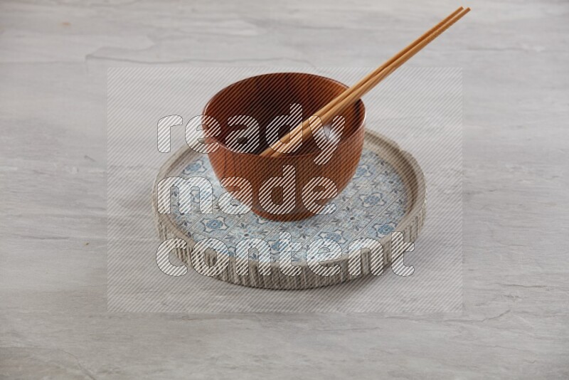 brown wood round bowl on top of multi color round ceramic plate and chopsticks, on grey textured countertop