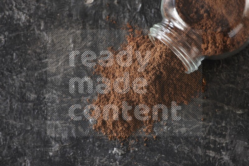 A flipped glass spice jar full of cloves powder and powder came out of it on textured black flooring