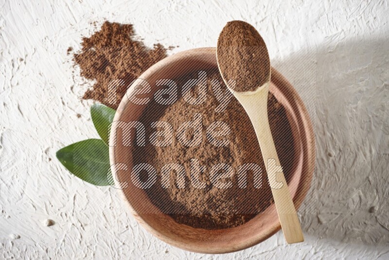 A wooden bowl and a wooden spoon full of cloves powder on a textured white flooring