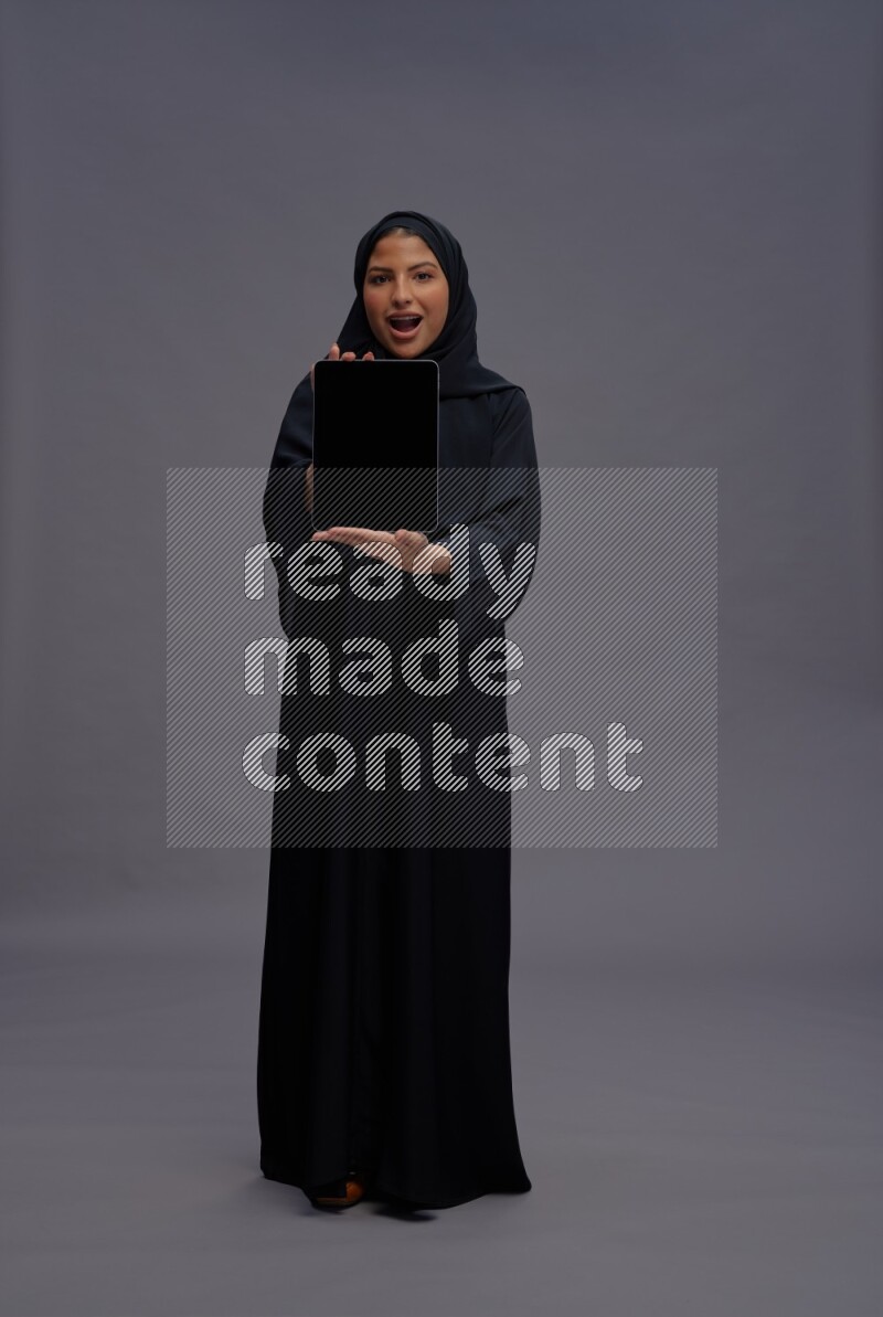 Saudi woman wearing Abaya standing showing tablet to camera on gray background