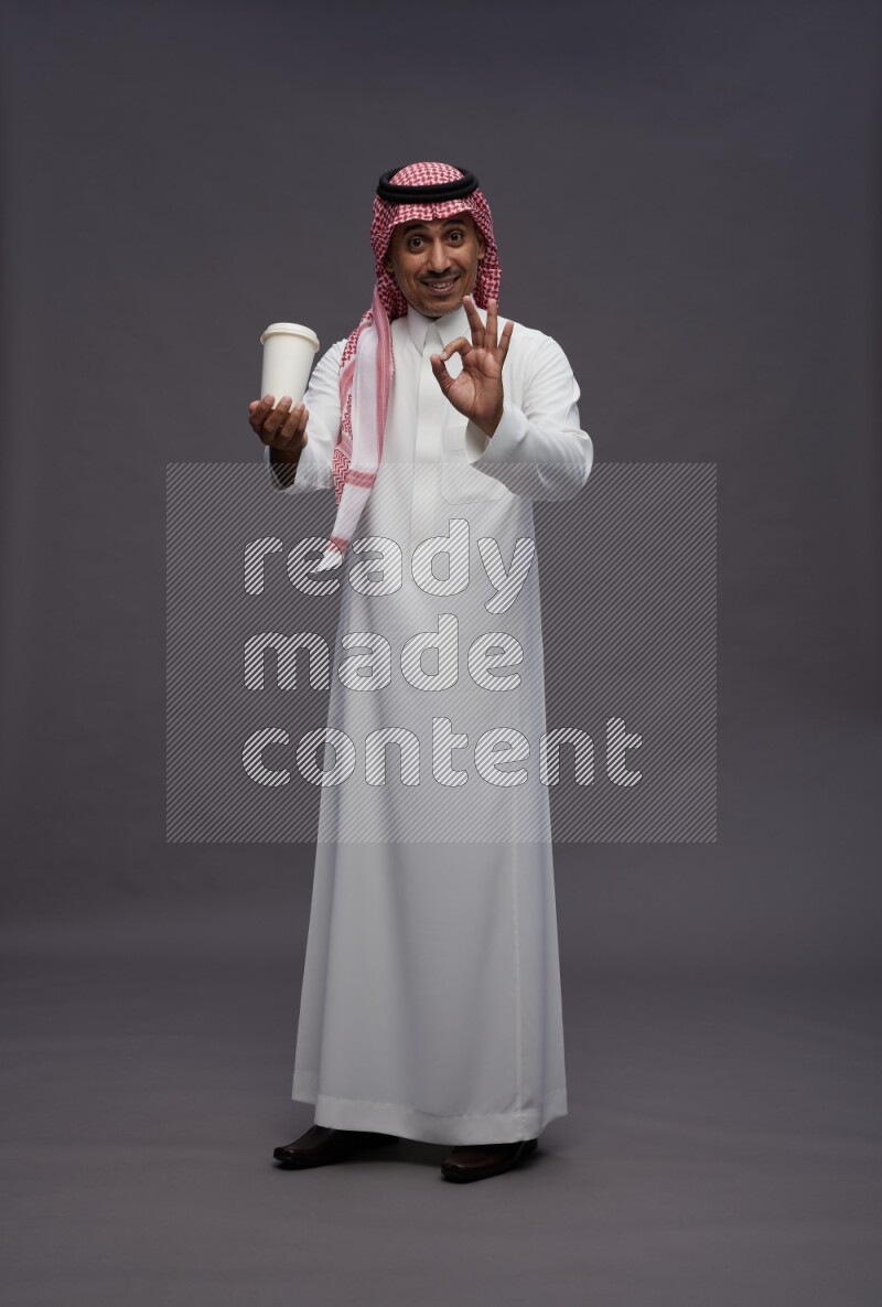 Saudi man wearing thob and shomag standing holding paper cup on gray background