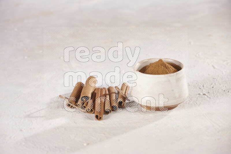 Cinnamon sticks stacked beside a beige bowl full of cinnamon powder on white background