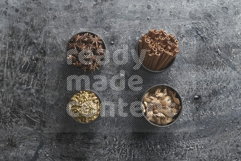 Ginger, Cardamom, Star anise and cinnamon sticks in 4 bowls on a textured black background