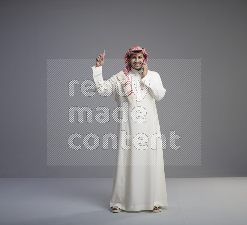 A Saudi man standing wearing thob and red shomag talking on phone on gray background