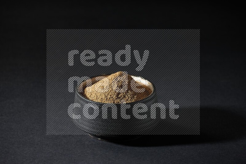 A black pottery bowl full of cumin powder on a black flooring