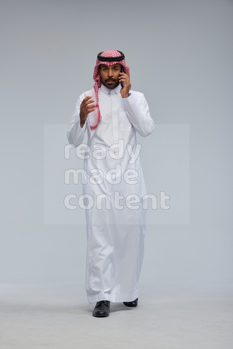 Saudi man Wearing Thob and shomag standing talking on phone on Gray background