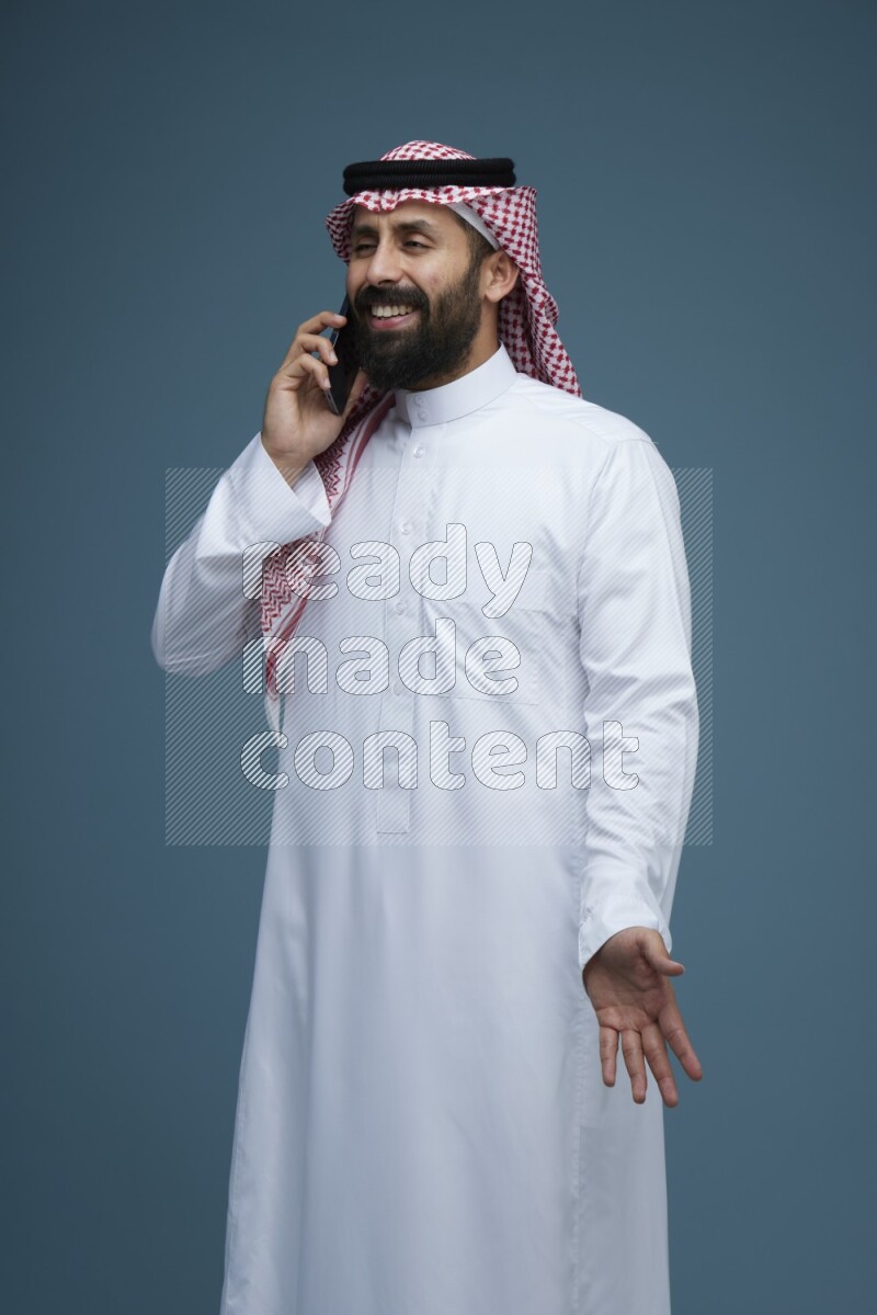 A man having a Call in a blue background wearing Saudi Thob with Shomag