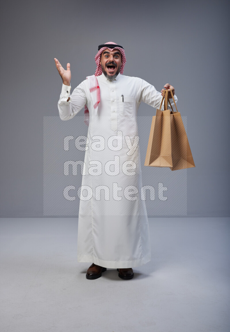 Saudi man Wearing Thob and red Shomag standing holding shopping bag on Gray background