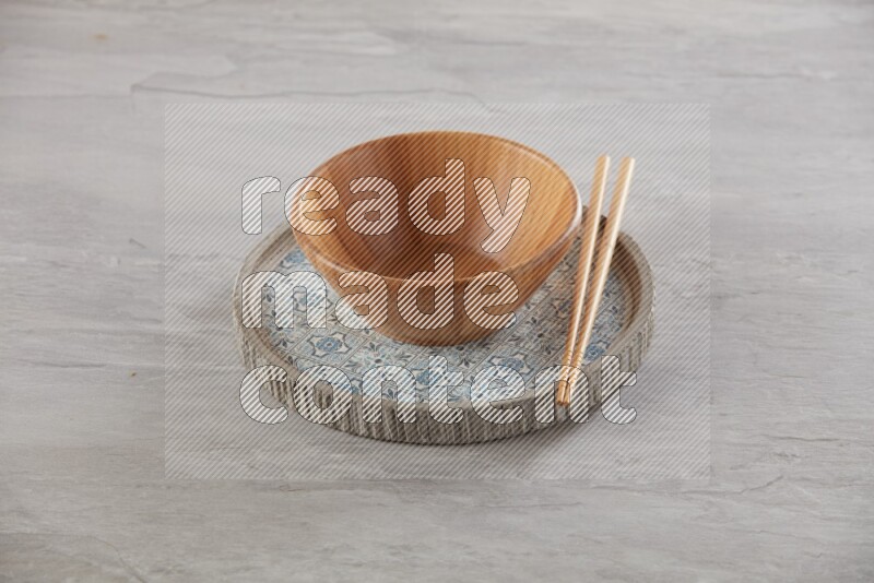 brown wood round bowl on top of multi color round ceramic plate and chopsticks, on grey textured countertop