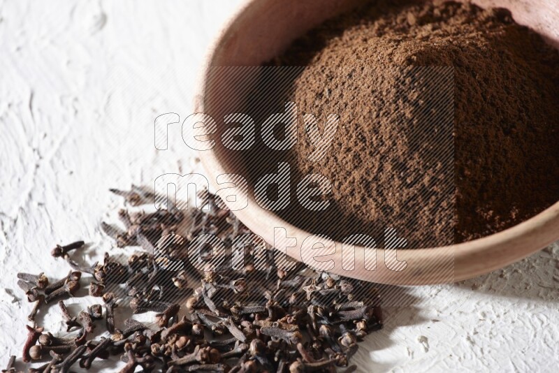 A wooden bowl full of cloves powder with whole cloves beside it on a textured white flooring