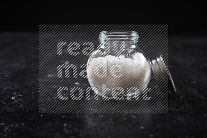 A glass jar full of coarse sea salt crystals on black background