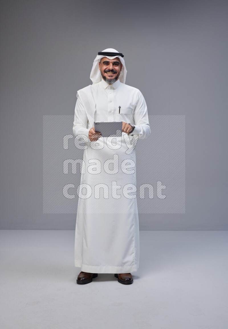 Saudi man Wearing Thob and white Shomag standing working on tablet on Gray background