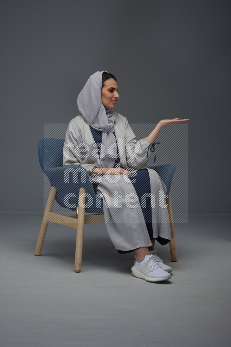A Saudi woman wearing a light gray Abaya and head scarf sitting on a dark grey chair while making multi hand gestures eye level on a grey background