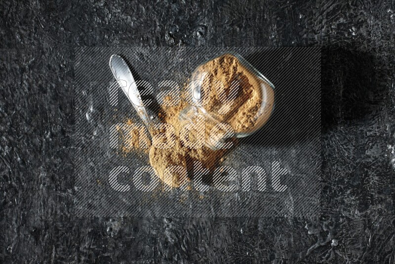 وعاء زجاجي للتوابل مقلوب وملعقة معدنية ممتلئان ببودرة بهار الفلفل الحلو والبودرة تناثرت من الوعاء على خلفية سوداء