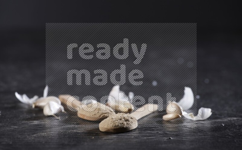 Two wooden spoons filled with garlic powder, surrounded by peeled garlic cloves and their skins on a textured black flooring