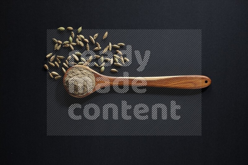 A wooden ladle full of cardamom powder and cardamom seeds beside it on black flooring