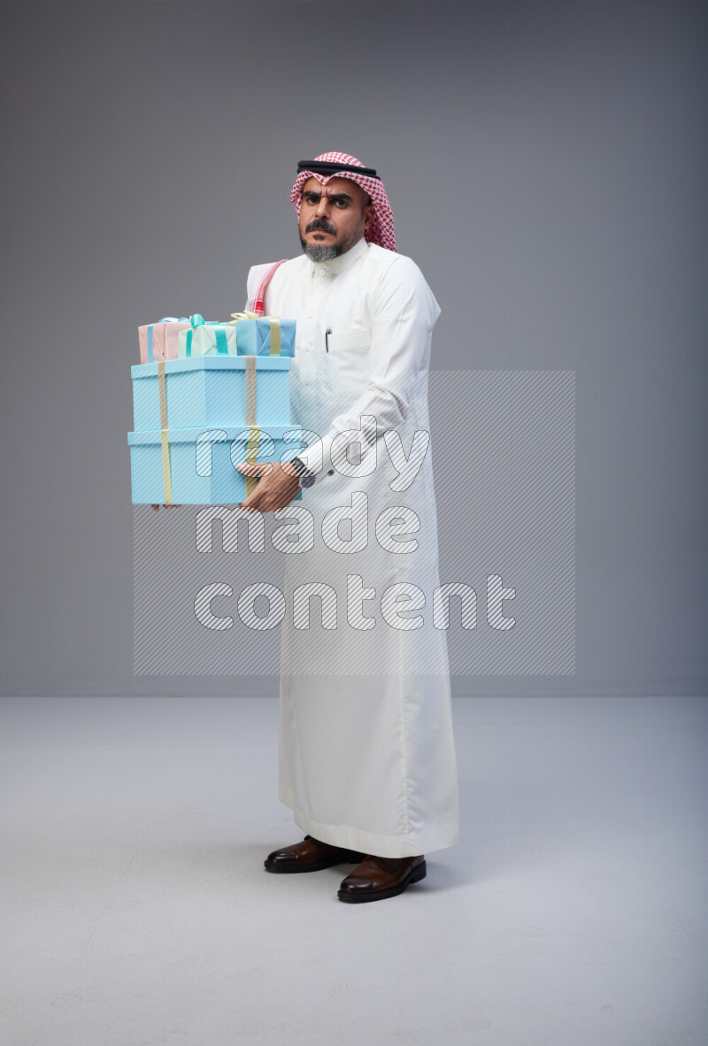 Saudi man Wearing Thob and red Shomag standing holding gift box on Gray background
