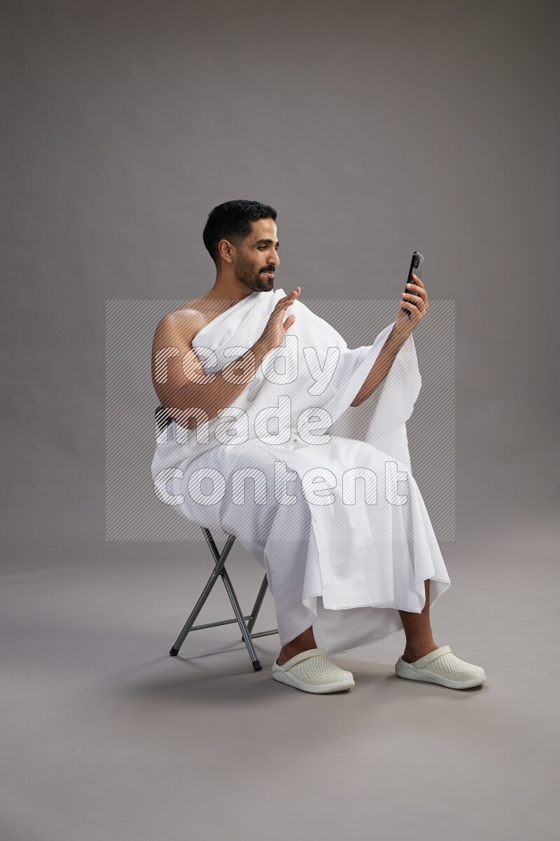 A man wearing Ehram sitting on chair taking selfie on gray background