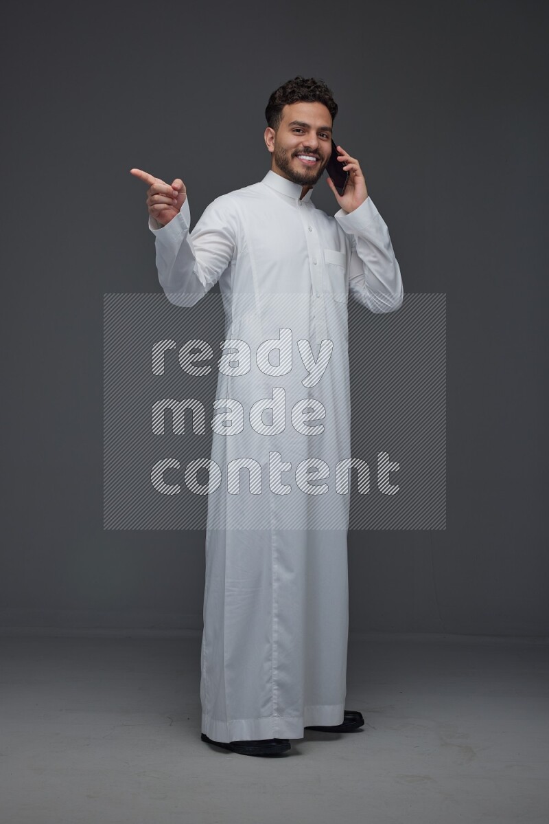 A Saudi man wearing Thobe and talking in the phone while standing and making different hand gestures eye level on a gray background
