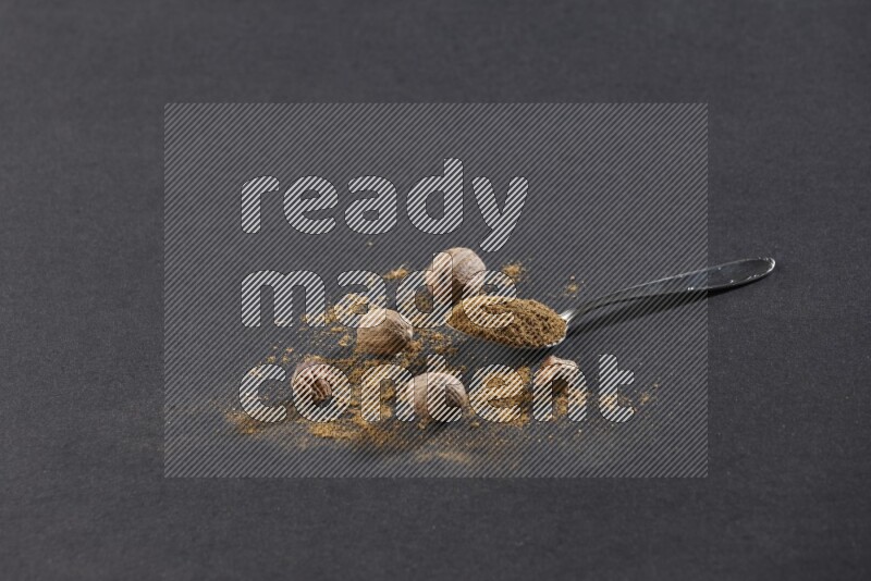 A metal spoon full of nutmeg powder with nutmeg powder and whole seeds beside it on a black flooring