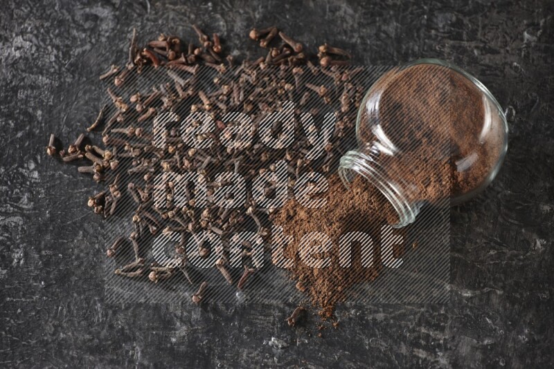 A flipped glass spice jar full of cloves powder and powder came out of it with cloves spread on textured black flooring