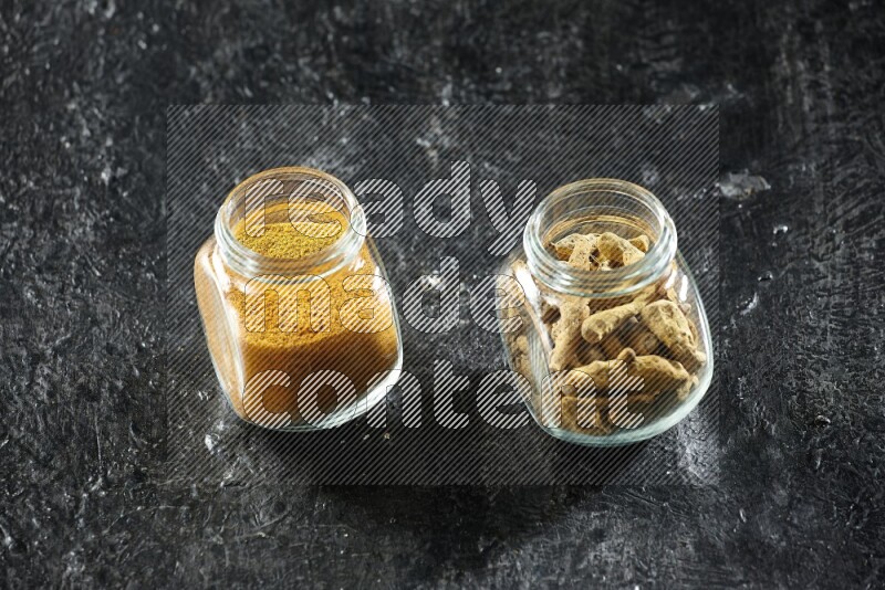 2 glass spice jars full of turmeric powder and dried whole fingers on textured black flooring