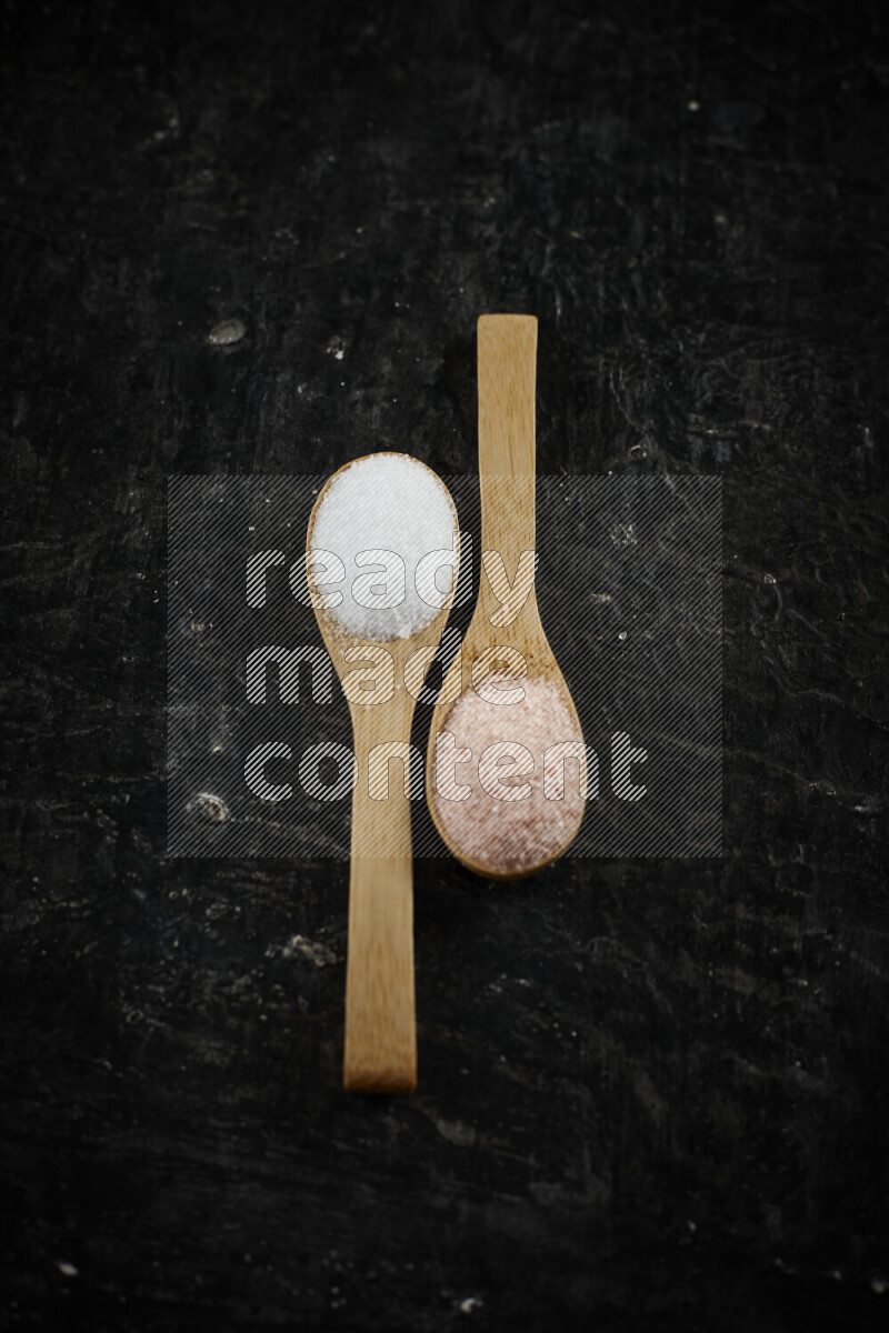 2 wooden spoons filled with fine and coarse salt on black background