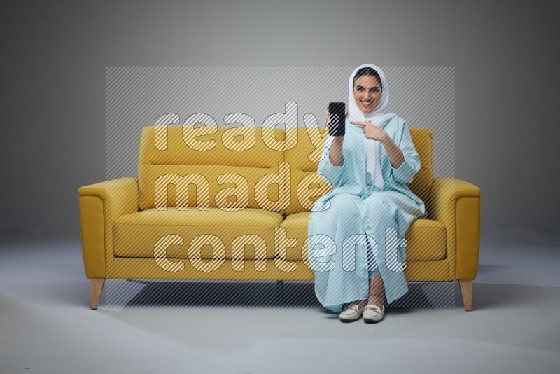 A Saudi woman wearing a light blue Abaya and white head scarf sitting on a yellow sofa and showing her phone's screen vertically and horizontally eye level on a grey background