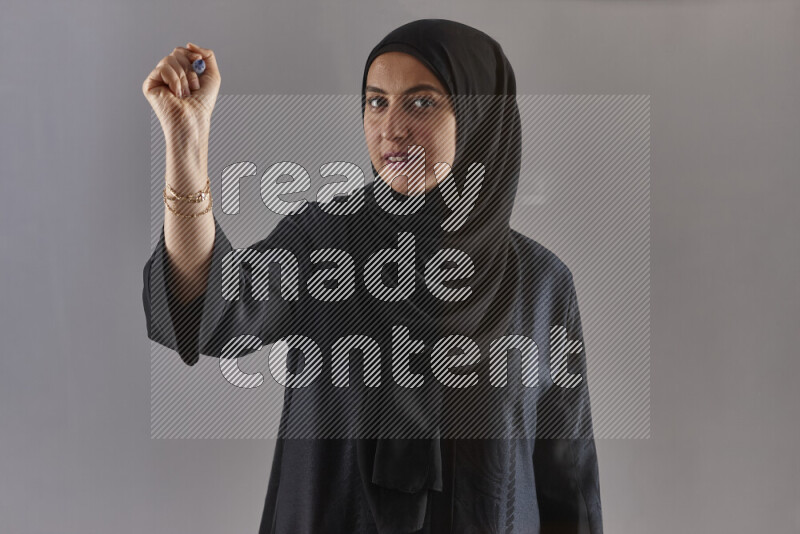 Woman in black abaya and hijab holding a marker pen to write on a transparent board with different reactions and expressions on grey background