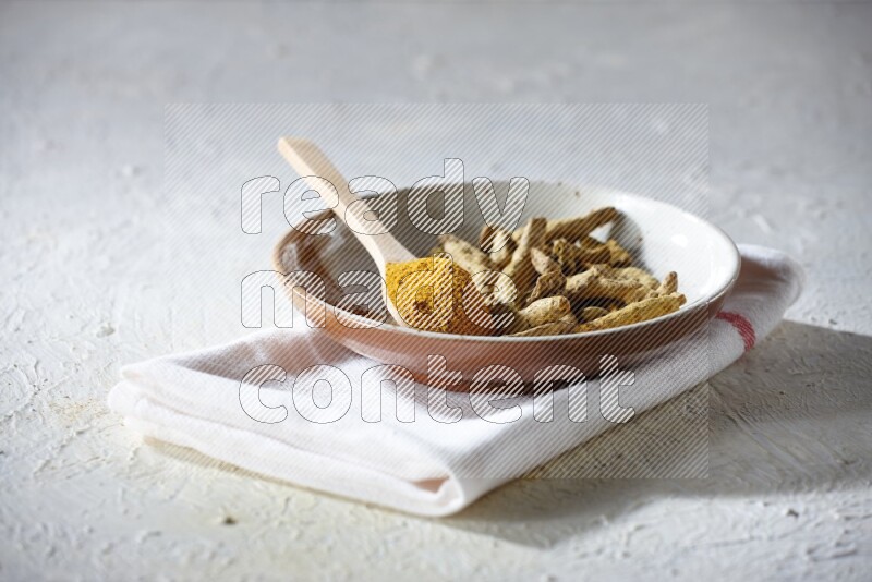 A plate filled with dried turmeric whole fingers and a wooden spoon full of turmeric powder on a textured white flooring