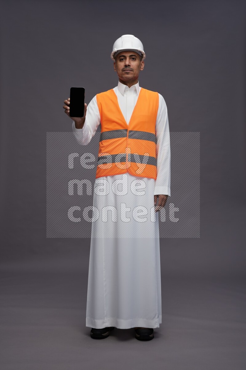 Saudi man wearing thob with engineer vest standing showing phone to camera on gray background