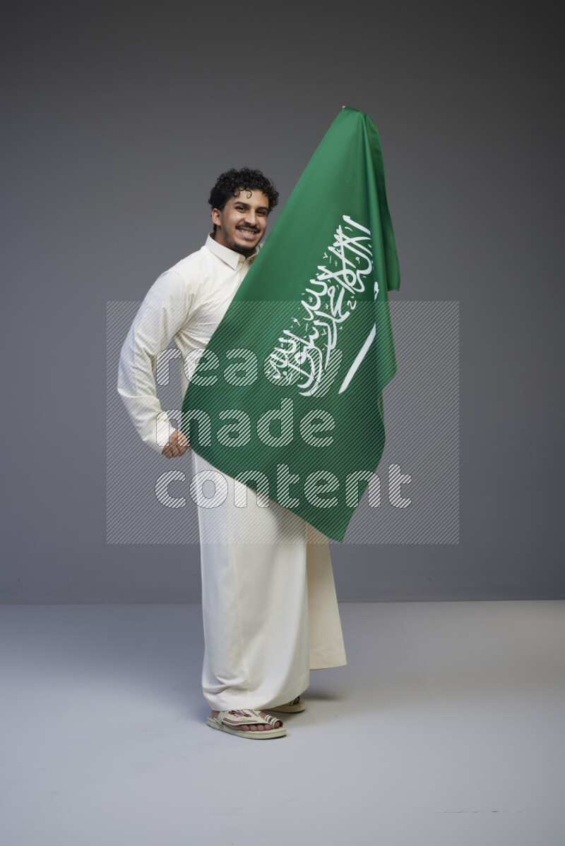A Saudi man standing wearing thob holding big Saudi flag on gray background