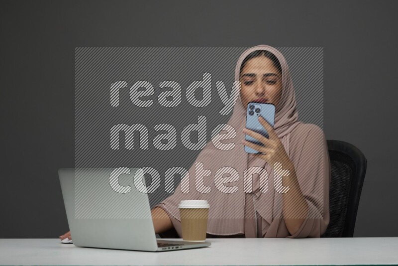 A Saudi woman Sitting on her desk Texting on a Gray Background wearing Brown Abaya with Hijab