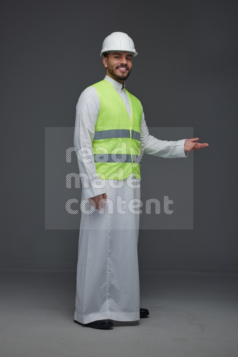 A Saudi man wearing Thobe with a yellow safety vest and white helmet standing and pointing different angles eye level on a gray background