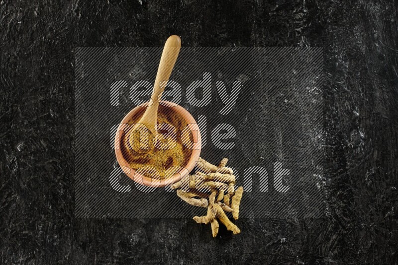 A wooden bowl and a wooden spoon full of turmeric powder with dried turmeric fingers on textured black flooring