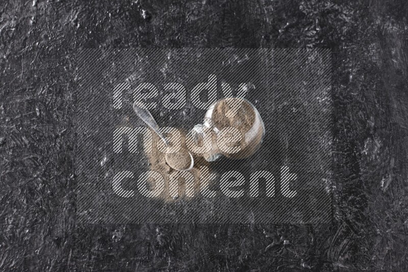 A flipped glass spice jar full of black pepper powder with a metal spoon full of powder on textured black flooring