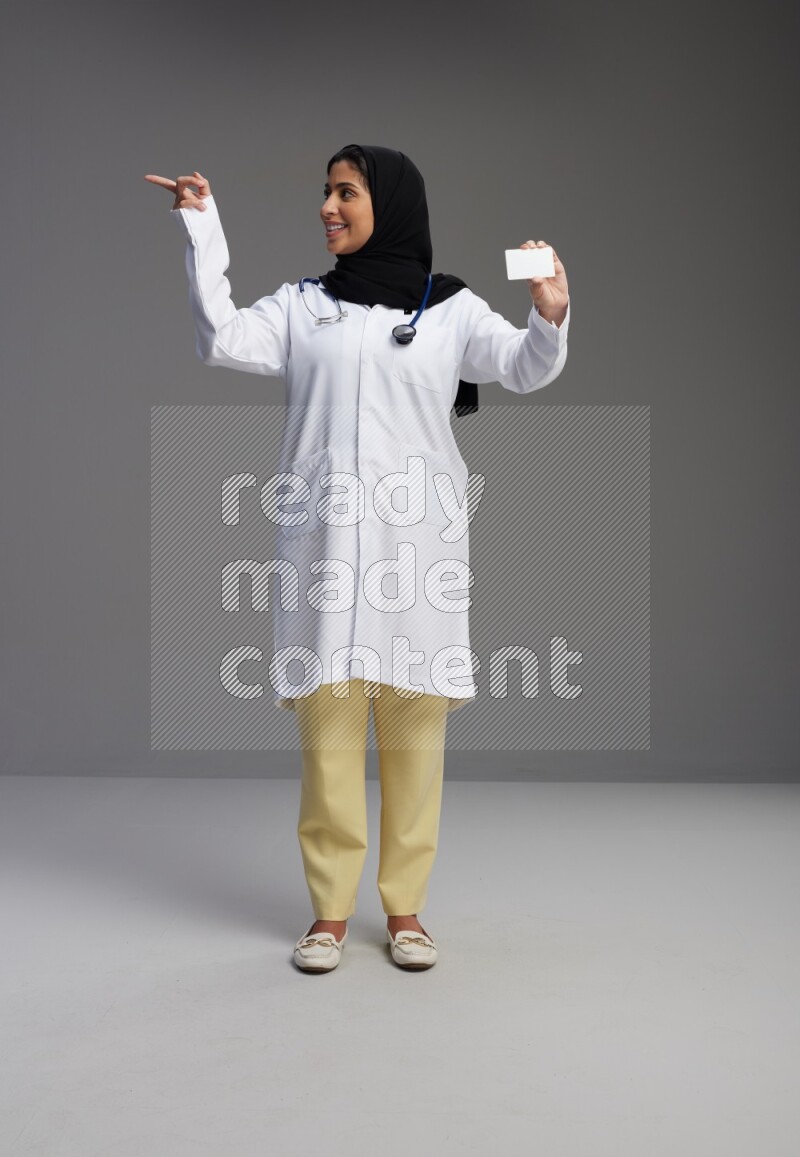 Saudi woman wearing lab coat with stethoscope standing holding ATM card on Gray background